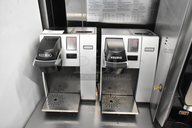 Two Keurig K150 countertop single cup coffee makers, silver and black, used, visible controls, sturdy drip trays.