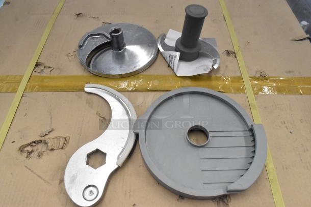 4 food processor accessories including S blade and slicing blade, metal and plastic, used condition, on cardboard surface.