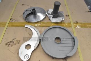 4 food processor accessories including S blade and slicing blade, metal and plastic, used condition, on cardboard surface.