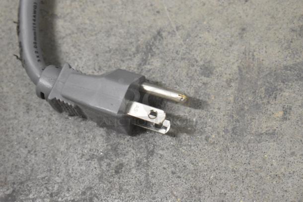 Gray power cord with a three-prong plug on a concrete surface, part of the Amana RCS10TS microwave.