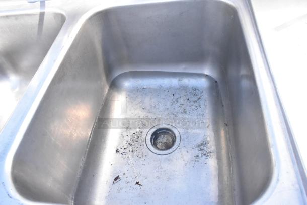 Stainless steel 2-bay sink with dual drain boards, faucet, and handles. Visible signs of wear, ideal for commercial use.