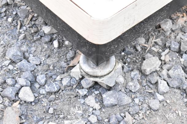 Metal display rack on casters, featuring a sturdy corner construction, shown on uneven rocky ground.