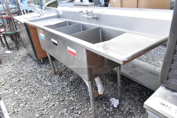 Stainless steel commercial 3 bay sink with dual drain boards, visible wear, color-coded labels, sturdy metal legs.