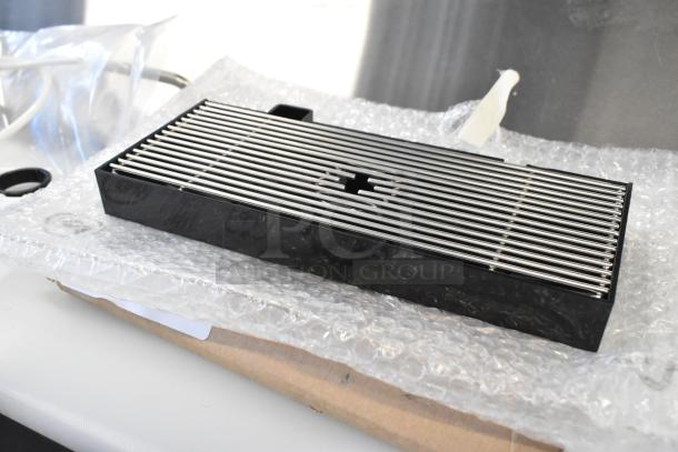 Brand new drip tray with minor scratches, metal grating, and black plastic frame, on bubble wrap for auction.