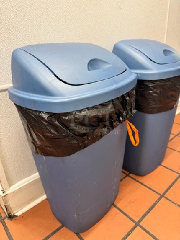 Blue swing top waste can, model BIDX2, with lid and black liner, in used condition on terracotta tile floor.