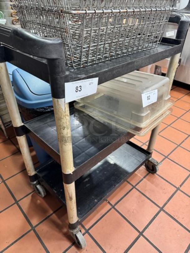 Three-tier bus cart with metal frame, plastic shelves, and caster wheels; visible wear, holds wire basket and clear bins, numbered tags.