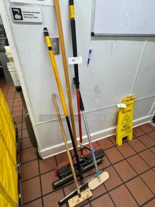 Assorted cleaning tools, including brooms and mops, leaning against a wall. Number tag 93 attached. Good condition.