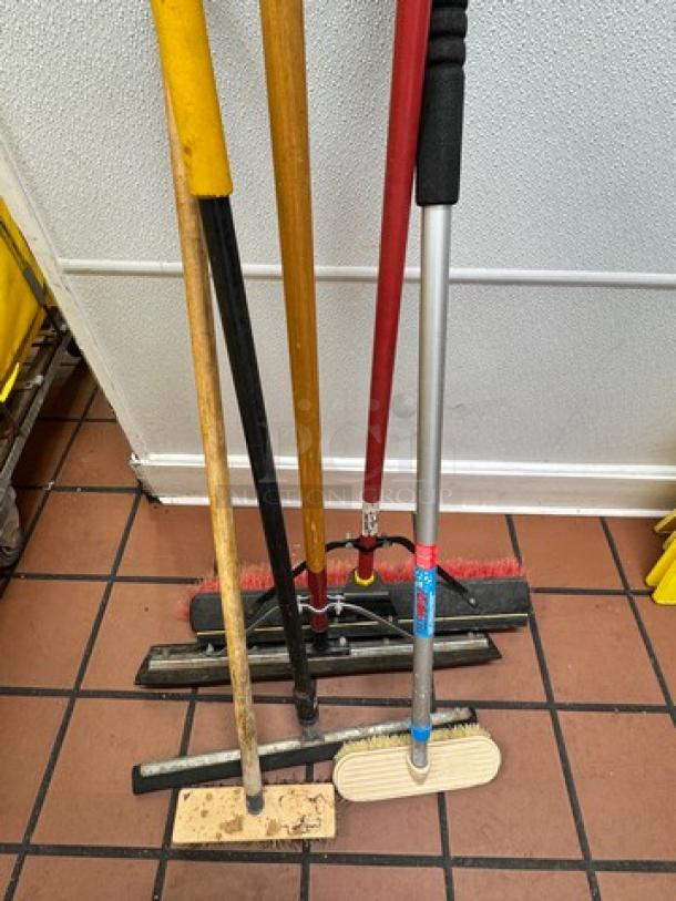 Various cleaning tools grouped together, including brooms and mops with different colored handles, in used condition.