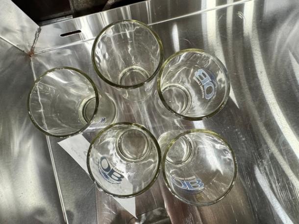 Lot of 5 clear glasses with "OB" logo, set on a reflective surface. Glasses appear in good condition.