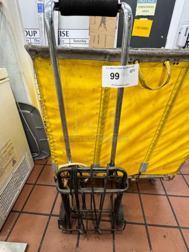 Rolling utility shopping cart with metal frame, black foam handle, and two wheels. Auction tag reads "99 QTY PCIAuction.com." Good condition.