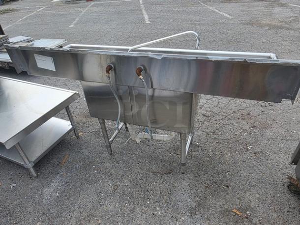Stainless steel two compartment sink with legs, visible piping, and faucet mounts. Condition appears used. Ideal for commercial use.