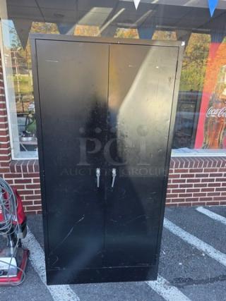 Black metal storage cabinet with dual doors and handles, showing wear and scratches.
