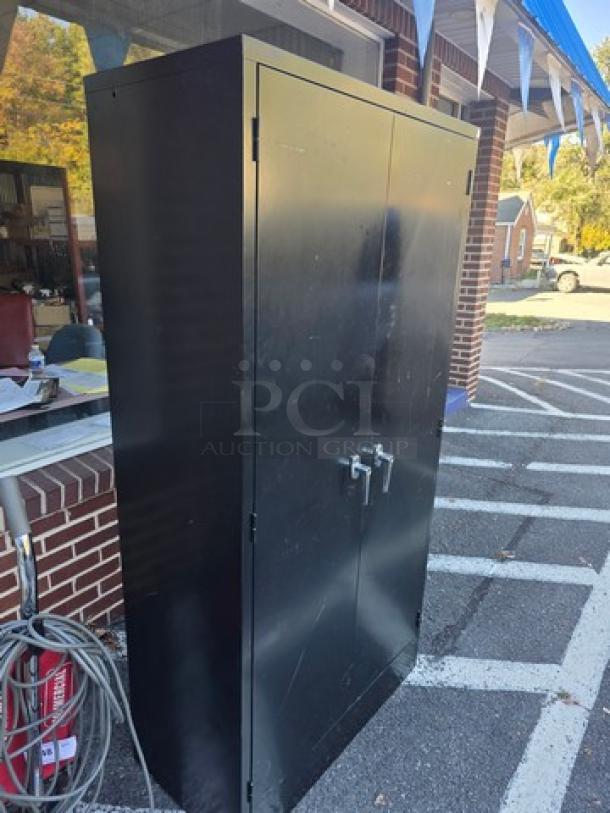 Black metal storage cabinet with double doors, sturdy construction, and handles. Visible scratches indicate light wear.