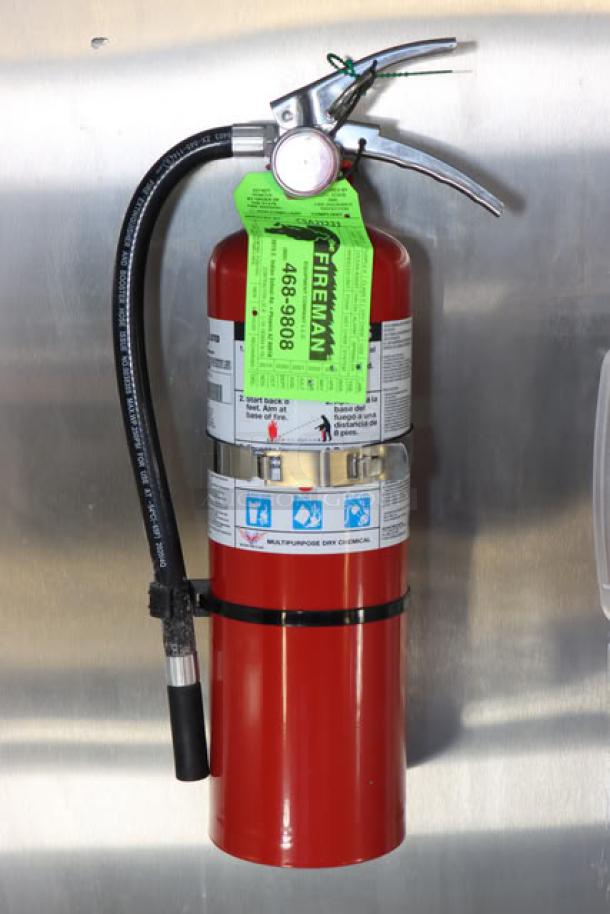 Red fire extinguisher mounted on stainless steel wall with visible green inspection tag labeled "FIREMAN 468-9808."