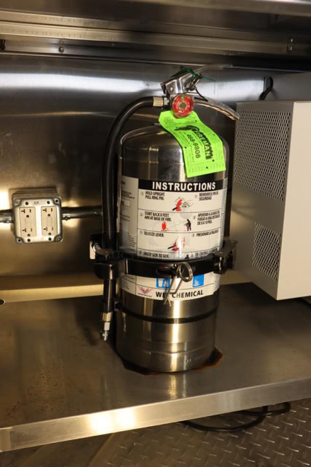 Stainless steel fire extinguisher mounted inside new Cruising Kitchens 24' food trailer, labeled with usage instructions.