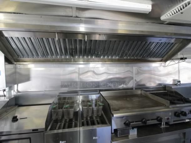 Interior view of a 2023 Cruising Kitchens 24 ft. Enterprise Food Trailer. Features deep fryers, griddle, range, and 12 ft. hood with ANSUL system. Stainless steel commercial-grade appliances and fixtures visible.