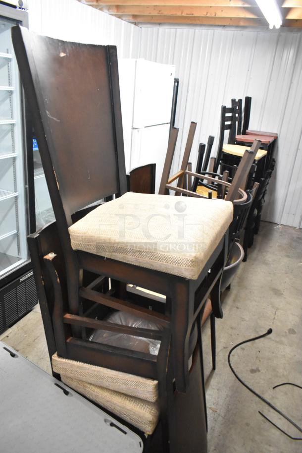 19 assorted chairs stacked in a storage area. Features various styles and finishes, some with fabric seats. Condition varies.