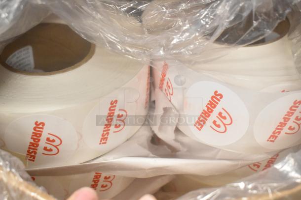 Rolls of "SURPRISE!" stickers in plastic wrap, part of a mixed auction lot. Stickers feature red text on round, white labels.