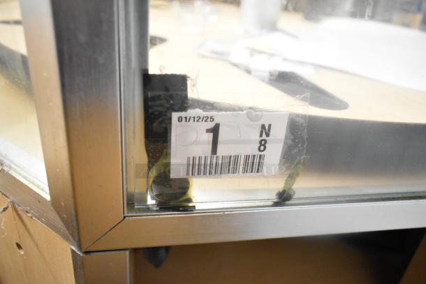 Merchandiser display case with metal frame and glass panels, featuring barcode sticker with serial 01/12/25, showing wear.