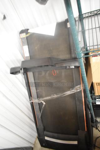 Folding treadmill in storage position, black with orange accents, appears in good condition. Packaging tape visible on frame.