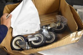 Lot of 4 commercial casters in a cardboard box, featuring metal frames and black rubber wheels, new condition.