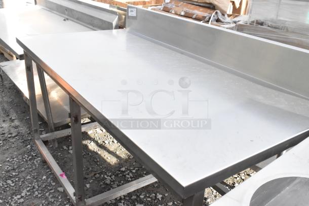 Win-holt stainless steel table with backsplash, in used condition. Features sturdy legs and smooth surface. Ideal for kitchens or workshops.