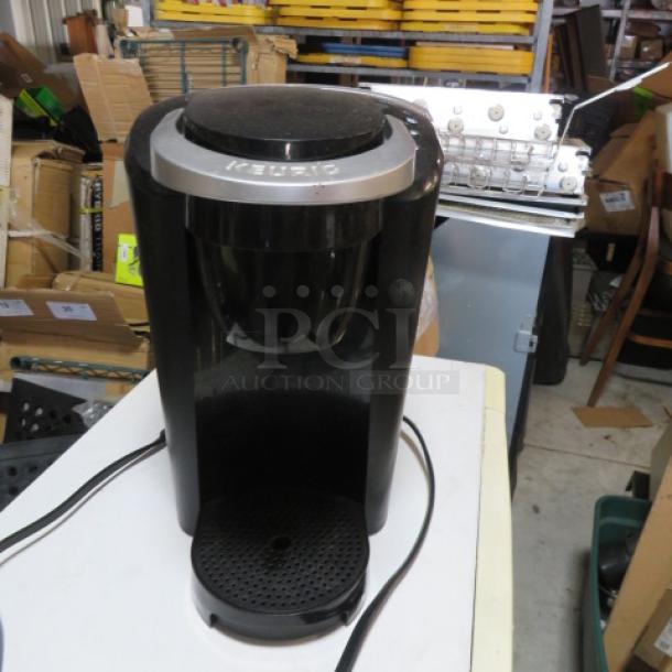 Black Keurig coffee brewer, pre-owned, slight scuffing, power cord attached. Keurig logo visible.