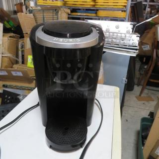Black Keurig coffee brewer, pre-owned, slight scuffing, power cord attached. Keurig logo visible.