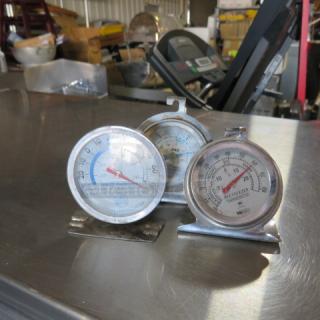 Three assorted metal thermometers on a metallic surface, each with temperature markings; used condition.