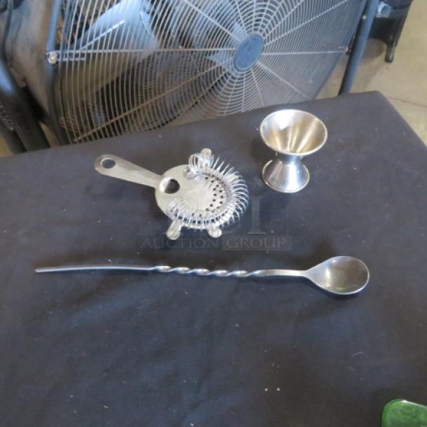 Stainless steel bar accessories set with cocktail strainer, jigger, and twisted mixing spoon on display.