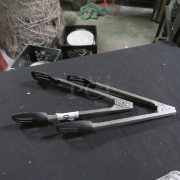 Stainless steel tongs with black grips, marked “2XBID,” on dark surface. Used condition, suitable for cooking or serving.