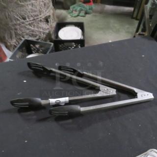Stainless steel tongs with black grips, marked “2XBID,” on dark surface. Used condition, suitable for cooking or serving.