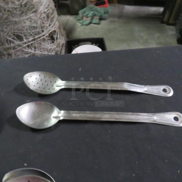 Two stainless steel spoons on a black surface; one slotted, both with elongated handles and hanging holes; used condition.