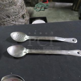 Two stainless steel spoons on a black surface; one slotted, both with elongated handles and hanging holes; used condition.