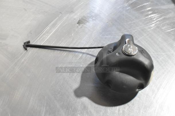 Black locking fuel cap with tether, featuring metallic key insert, placed on metallic surface.