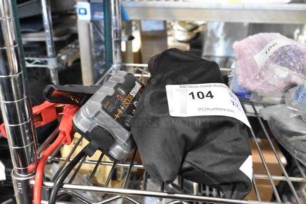 NOCO GB40 Genius Boost jump starter with clamps, black carrying bag labeled "104," on a metal shelf.