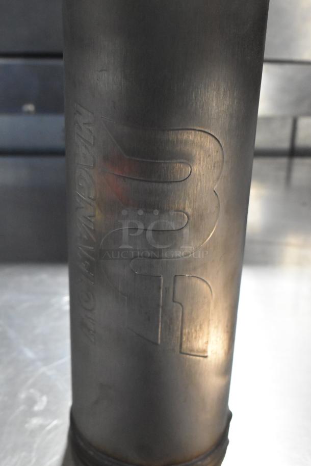 Magnavox MF exhaust component, metallic finish, engraved brand logo, showing slight wear, paired with Noco GB40 Genius Boost.