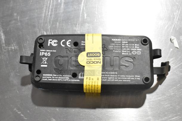 Noco GB40 Genius Boost jump starter, model GB40, 12V, IP65, 24Wh, with caution labels, on metal surface.