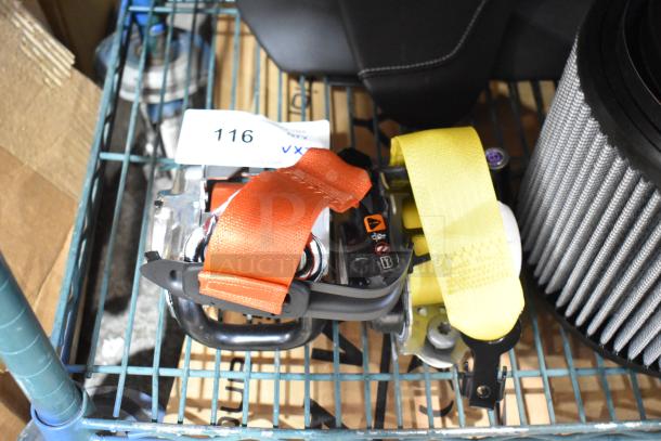 Two brightly colored seat belts displayed on a metal cart. Orange and yellow straps with metallic components. Auction tag labeled "116 VX" visible.