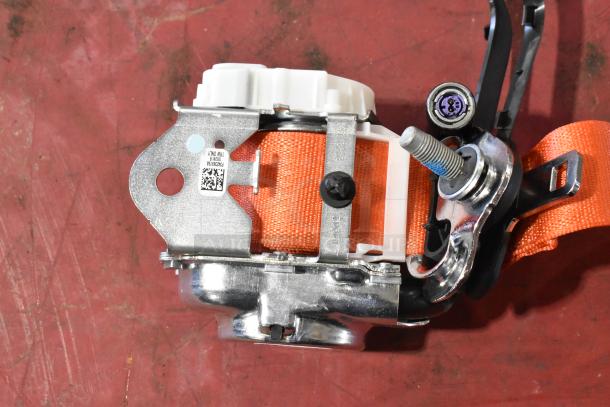 Seat belt mechanism with orange strap, chrome fittings, and visible QR code on metal plate, indicating part details.