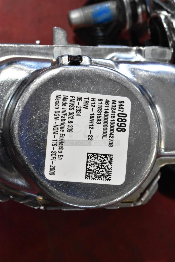 Metallic automotive component with a label showing model number, manufacturing details, and a QR code, part of a seat belt system.