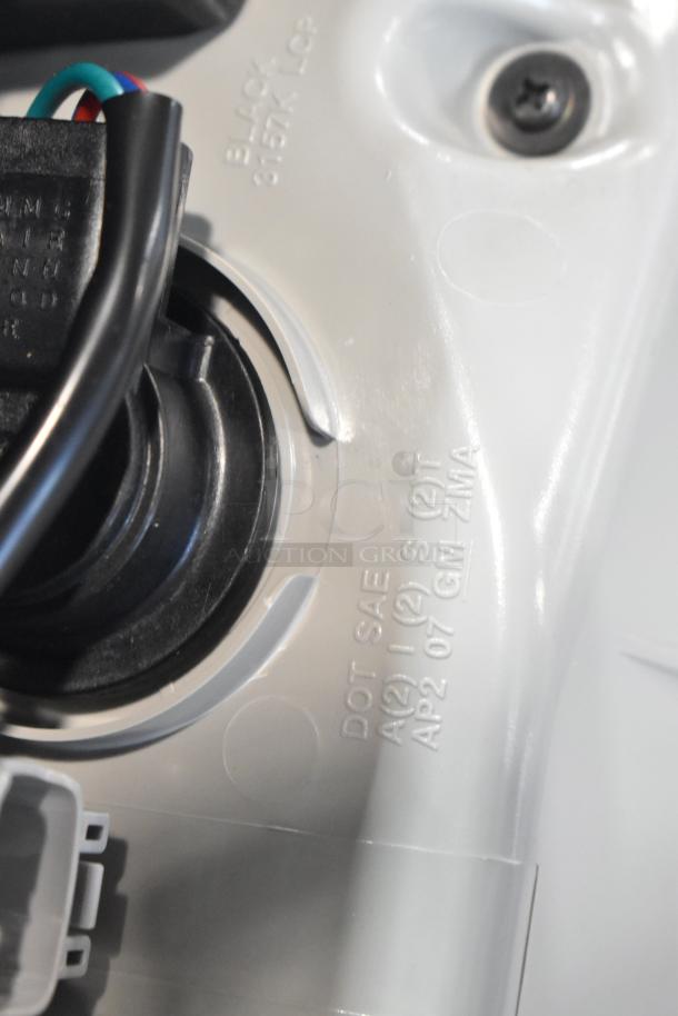 Close-up of a white automotive part labeled "DOT SAE A(2) I (2) S (2)T AP2 07 GM ZMA" with visible wiring and connector.