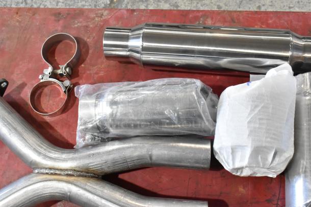 Assorted exhaust pieces with clamps and connectors, varying materials and conditions, some wrapped in plastic.