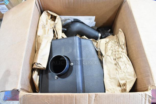 Box containing automotive parts, including a black air intake component, in new condition with packing paper.
