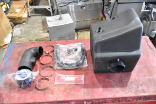 Box of parts, including a black plastic housing, elbow pipe, hose clamps, and sealed components. New condition.