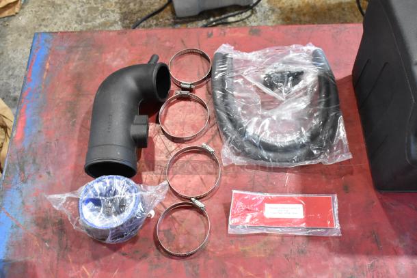 Box of parts includes black elbow pipe, four metal clamps, black hose, and packaged item with labeled tag on red surface.