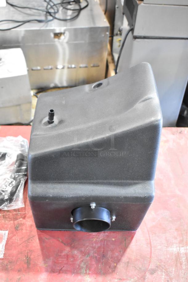 Black plastic part from a box of parts, featuring a pipe connector and vent opening, in used condition on a red surface.