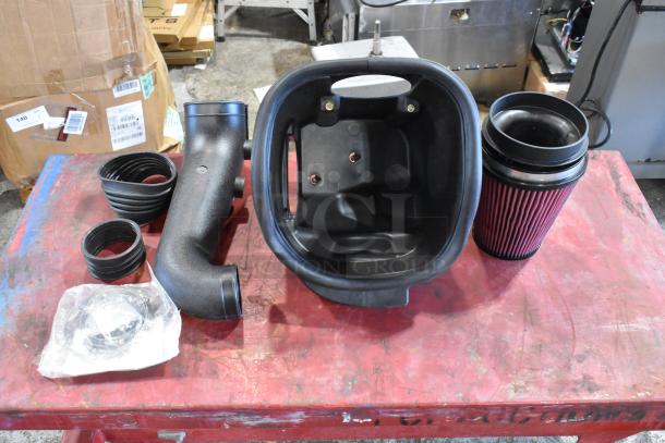 Box of automotive parts, including air filter, hoses, and plastic housing, shown on a red workbench. New condition.