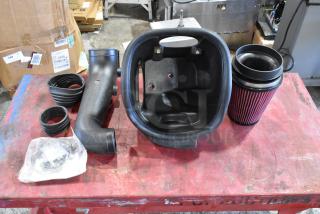Box of automotive parts, including air filter, hoses, and plastic housing, shown on a red workbench. New condition.