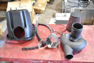 Box of automotive parts including a cold air intake kit with filter, hoses, clamps, and housing. Parts appear new.